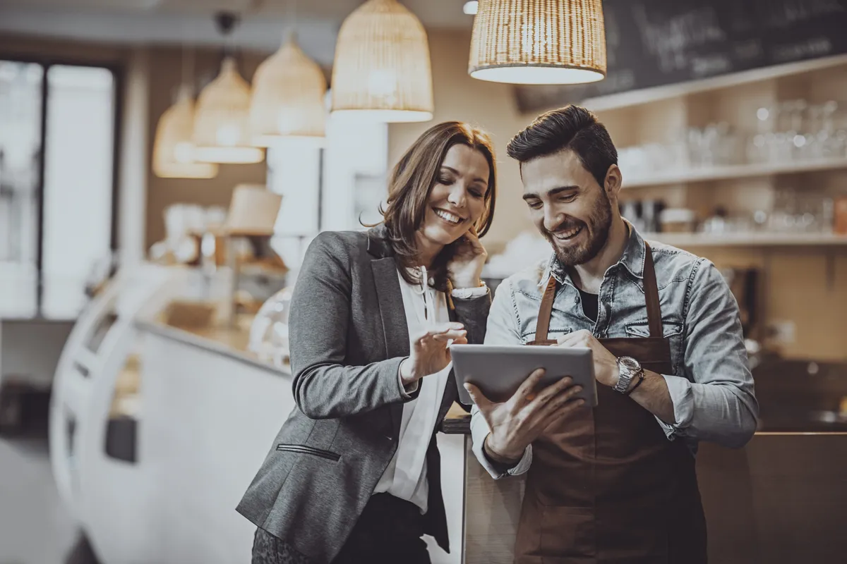 A coffee shop owner and a colleague looking at a tablet together behind the counter.