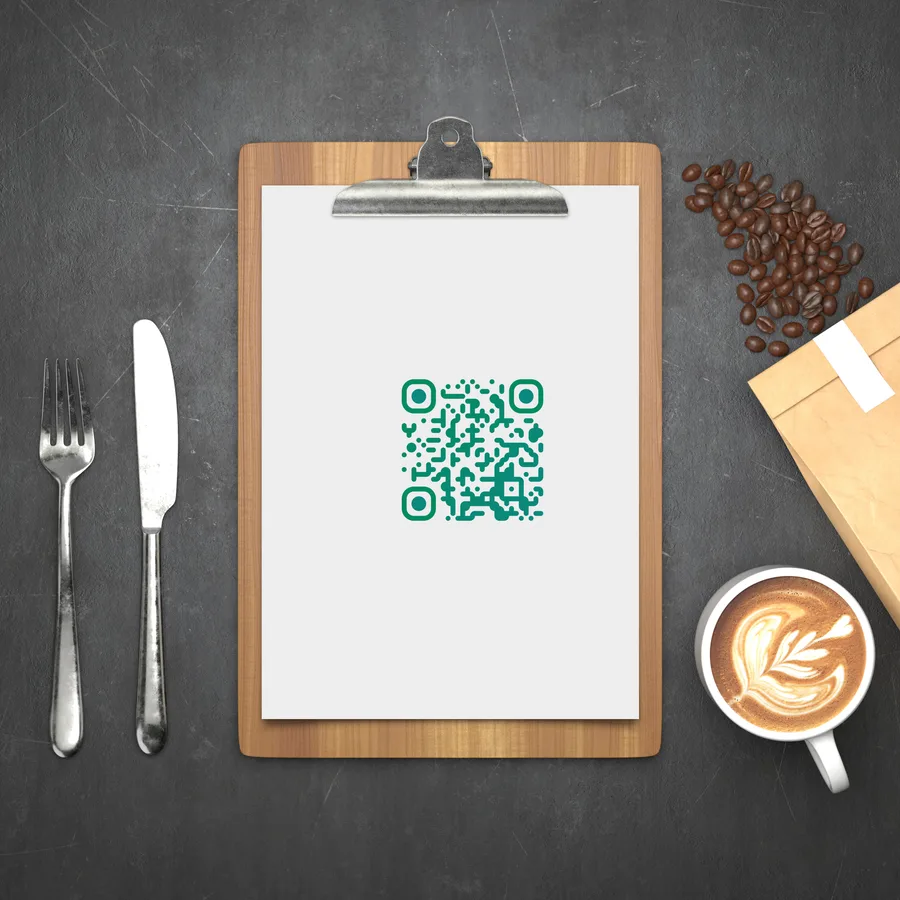 A QR code printed on a restaurant menu clipboard beside cutlery and a coffee.